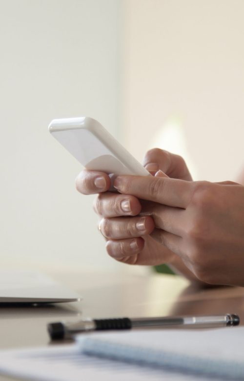Close up image of modern cellphone in womans hand at desk with laptop. Office worker boring at workplace and playing games, writing in social networks. Businesswoman looking important contact on phone